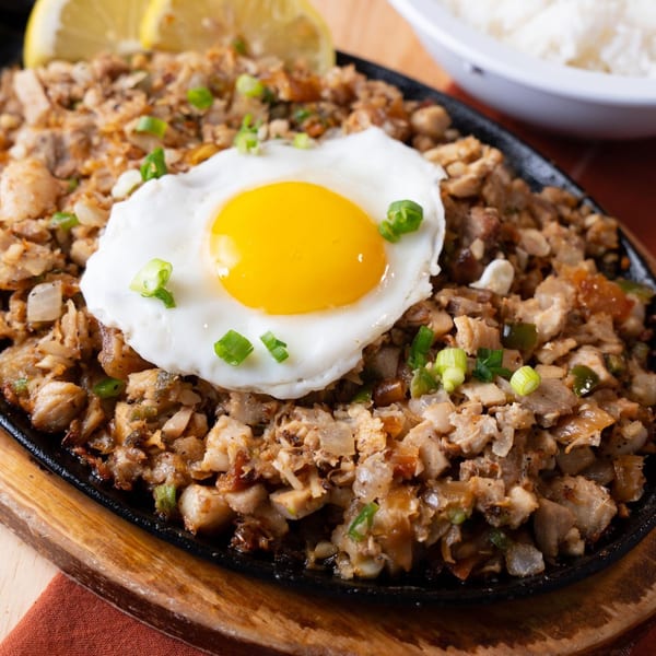 Filipino sisig with a fried egg and green onions on a sizzling plate, with lemon and rice.