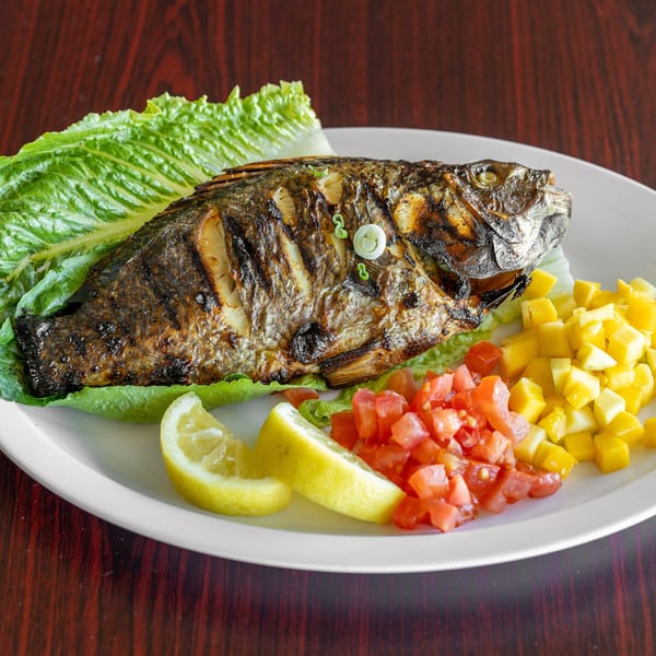 Grilled whole fish, garnished with lettuce, lemon wedges, diced tomatoes, and mangoes on a white plate.